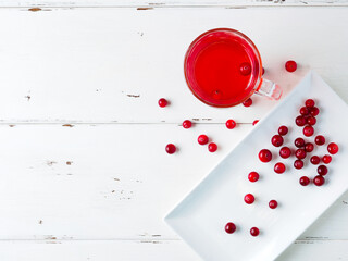 Selective focus on cranberries in a fresh drink in a glass cup on a white wooden background. Fresh ripe berries are scattered on a white rectangular ceramic plate. Copy space. Flat lay.