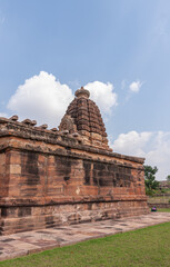 Aihole, Karnataka, India - November 7, 2013: Huchchimalli Gudi or Temple. Side red stone wall with sculpted vimanam towering over it under blue cloudscape. Green grass in front.
