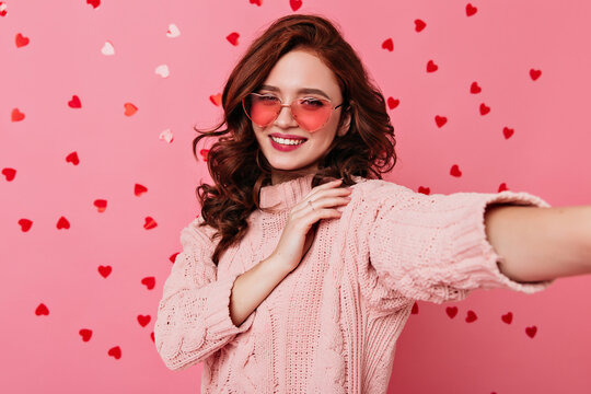 Romantic girl in glasses posing in valentine's day. Appealing young lady making selfie on pink background.