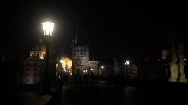 
street lights and statues on the stone Charles Bridge at night and silhouettes of walking people on the bridge at night in the center of Prague