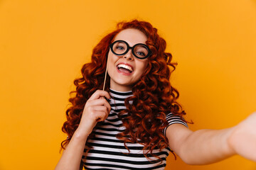 Naughty young woman in striped T-shirt poses with toy glasses and takes selfie in orange studio