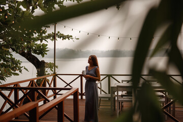 Woman enjoying hot drink on a sunset near the lake, stand on wooen pier. She wear long grey dress