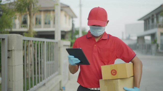 A male delivery worker wearing a red uniform Looking for a home that is currently shipping, search for the house number from the tablet you hold.New normal Concept.