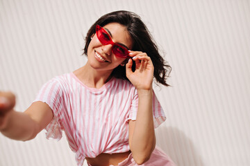 Blissful brunette woman taking selfie with smile. Well-dressed girl in pink sunglasses laughing at camera.