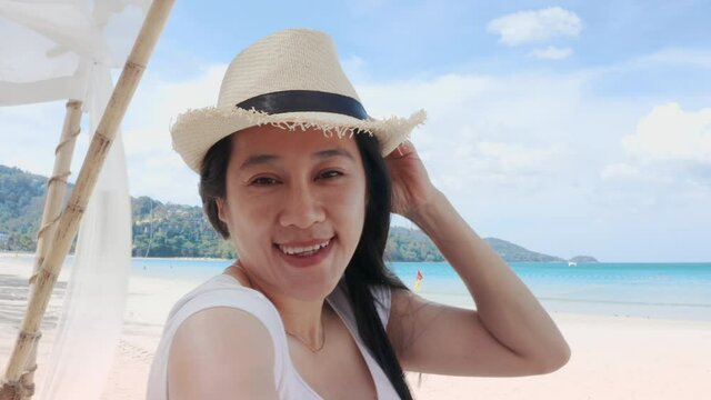 4K A happy carefree female tourist wear straw hat is making selfie or video call with phone to a friends or family on a desert offshore seaside in a sunny day during vacation trip.
