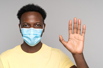 Young African American man wearing yellow t-shirt and protective face mask to safe from corona virus Covid-19 showing and rising palm up isolated on studio grey background. Copy space. 