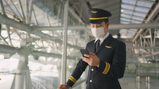 Asian young pilot wearing face mask, using mobile phone in terminal.