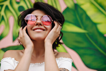 Front view of amazing girl in pink round glasses. Laughing female model posing near green graffiti.