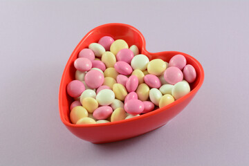 Pink, white and yellow Valentine's Day candy coated chocolate snack in red heart shaped bowl on lavender background