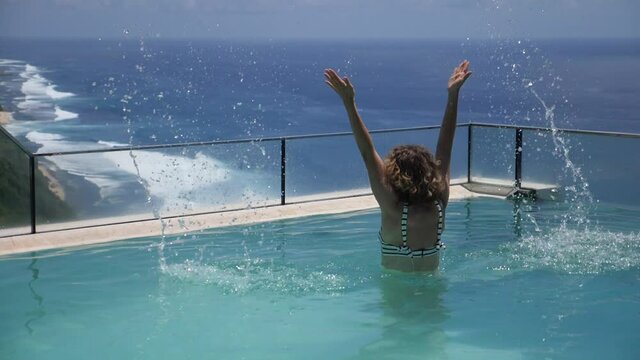 Living the best life. Woman jumps out of an infinity pool and splashes water. Ocean and the mountains on the background