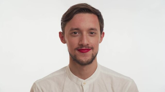 Young European confident brunette handsome male with brown beard, red lipstick wear shirt, smile, flirt, put hand to mouth and blow kiss on white background. Portrait metrosexual or gay man close up.