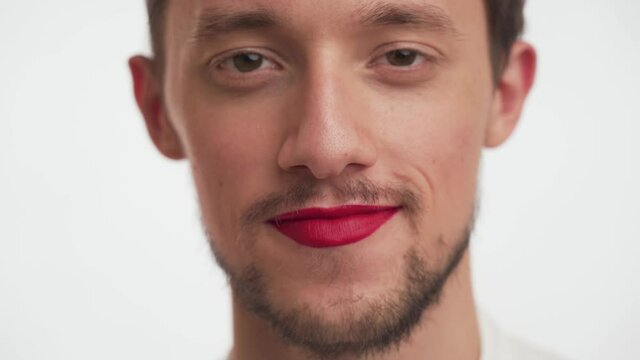 Close up portrait of male face with makeup. Young cute Caucasian man with beard, moustache, brown eyes painted lips by red color lipstick look at camera, smile on white background. Transvestite guy.