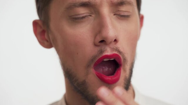 One bored Caucasian handsome brunette man with beard, moustache, painted lips by red lipstick opens his mouth wide, yawns on white background. Concept of boredom of beauty blogs. Close up male face