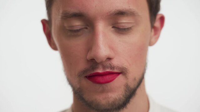 Handsome metrosexual Caucasian man with dark beard, brown eyes wear red lipstick on lips on white background close up. Portrait of one surprised guy, ореns mouth wide, covers with hand, stare camera.