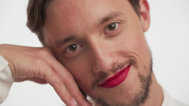 Handsome young brunette bearded guy with painted lips by red lipstick wear shirt, gently strokes soft skin of face with hand isolated on white background close up. Portrait of metrosexual or gay man.