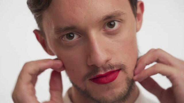 Beautiful Caucasian young brunette beauty man with beard painted lips in red lipstick, gently strokes soft skin of face with hand isolated on white background close-up. Portrait of transvestite guy.