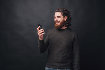 A cheerful young man is texting with someone smiling at the phone near a black wall .