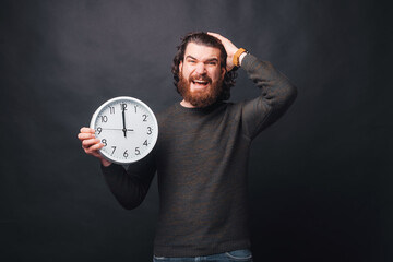 A young bearded man is looking stressed at the camera holding a wall clock .