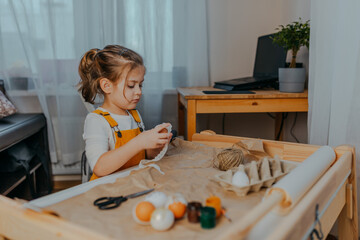 Baby girl decorating easter eggs on white table. Kid decorating eggs with kraft paper, lace and...