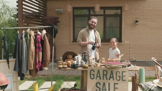  Handheld dolly-in shot of bearded father and little daughter standing behind table and chatting while preparing for garage sale in their front yard on summer day, then smiling for camera