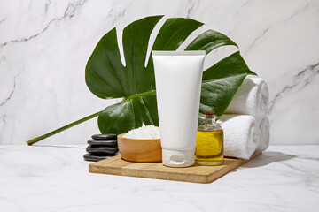 White cosmetic tube with spa element and towel with monstera leaf on marble background. Blank label for branding mock-up. Natural beauty product concept. 