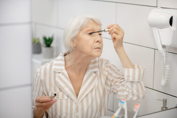 Gray-haired woman in pajama doing make up in the bathroom