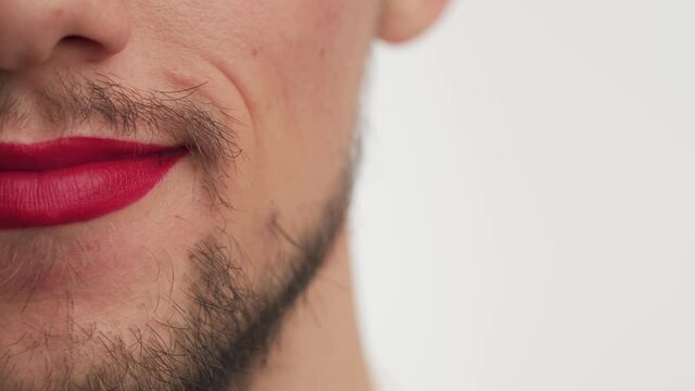 Close up view of single man mouth wear makeup – red lipstick on lips. Serious fashion Caucasian metrosexual or gay boy with black beard, moustache smiles isolated on white background. Copy space.