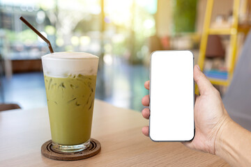 Iced green tea glass on wooden table on blurred coffee shop background. with men Hand holding new modern smartphone mobile phone with empty white display screen.