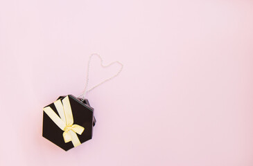 One dark black gift box with luxury gold bow with chain lined in the shape of a heart on pink background. Horizontal with copy space. Flat lay