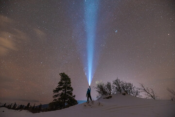 A person standing on a snow covered ridge in northern Canada holding a flashlight straight up with starry, star covered sky above at night time in wilderness area. 