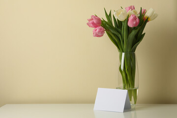 Vase with tulips and blank card against beige background, space for text