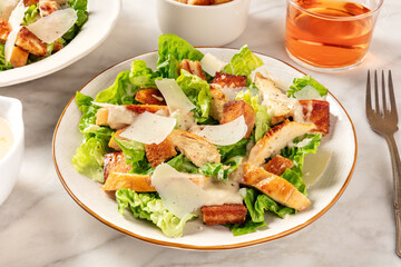 Caesar salad. Grilled chicken breast slices, green romaine salad leaves, croutons and parmesan, the classic recipe on a white marble background