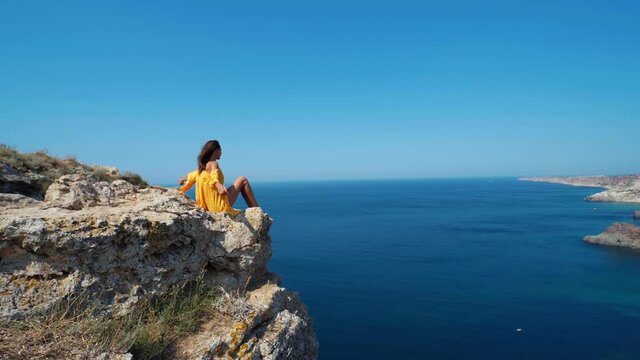 joyful taned woman tourist sitting on cliff top in front of amazing seascape, bright yellow dress and hair blowing in the wind. girl enjoying freedom of traveling and beautiful view