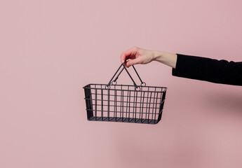 Female hand hold black metal supermarket busket on pink background