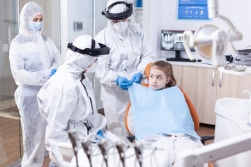 Little girl looking at dentist while assistant is putting her a bib before dental examination uring...