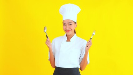 Lively and positive female chef holding fork and spoon posing on yellow studio background 