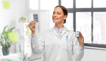 medicine, profession and healthcare concept - happy smiling female doctor comparing pills in jar and pack over medical office at hospital on background