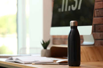Modern black thermos bottle on wooden desk at workplace. Space for text - obrazy, fototapety, plakaty