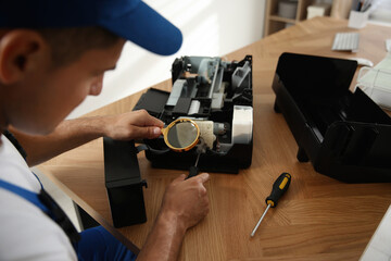 Repairman with magnifying glass and screwdriver fixing modern printer indoors, closeup