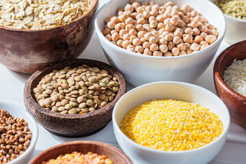 Assorted dry cereals, grains and legumes in white and wooden bowls, white marble background. Vegan protein concept.