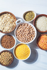 Assorted dry cereals, grains and legumes in white and wooden bowls, white marble background. Vegan protein concept.