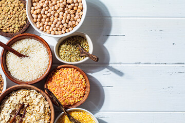 Assorted dry cereals, grains and legumes in white and wooden bowls, white wooden background, top view, copy space.  Vegan protein concept.