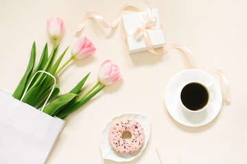 Morning cup of coffee, donut, gift or present box and spring tulip flowers on beige background. Beautiful breakfast for Women day, Mother day, Valentine's Day. Flat lay.