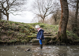 Excited happy young girl in wellinton boots walking through autumn countryside river stream splashing in water exploring the great outdoors nature park with bobble hat and blue coat in winter