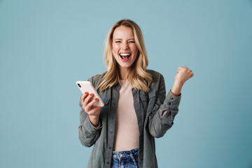 Delighted blonde girl making winner gesture and using mobile phone