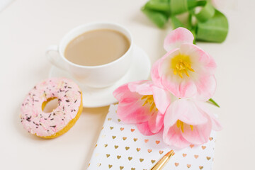 Spring breakfast for a blogger or office worker: a cup of coffee, a donat, a bouquet of pink tulips, a notebook with a pen
