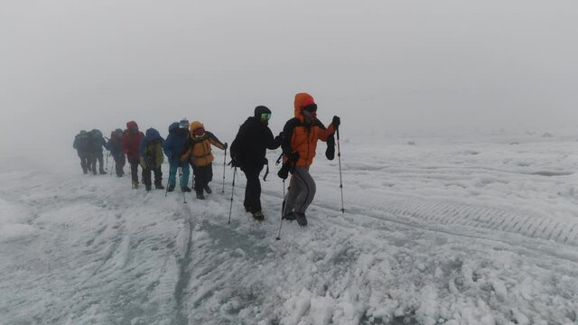Mountaineering activity on the steep mountain slope. Clip. Group of extreme tourists walking one by one on icy surface of a hill in foggy cold weather.