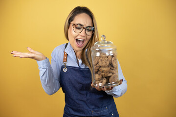 Young hispanic woman wearing baker uniform looking at a cookies jar confused with arms and hands...