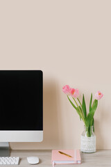 A computer monitor, a bouquet of pink tulips, and a notebook with a pen on a light wooden table against a beige wall. The workplace of a freelancer or student