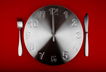 Lunch time. Metal clock with silver cutlery on the side. Studio photo isolated on red background. Selective focus on object.